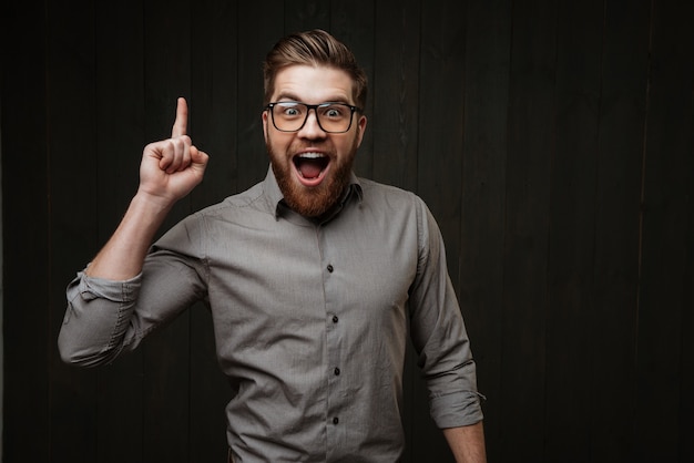 Retrato de un hombre barbudo feliz emocionado en anteojos apuntando con el dedo hacia arriba aislado en la superficie de madera negra