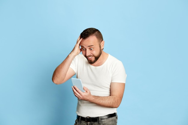 Retrato de un hombre barbudo feliz y emocionado con una camiseta blanca mirando emocionalmente el teléfono móvil con una cara positiva impactada contra el fondo azul Emociones humanas ventas compras concepto de apuestas Anuncio