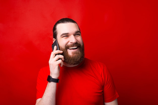 Retrato de hombre barbudo feliz hablando por teléfono inteligente sobre pared roja