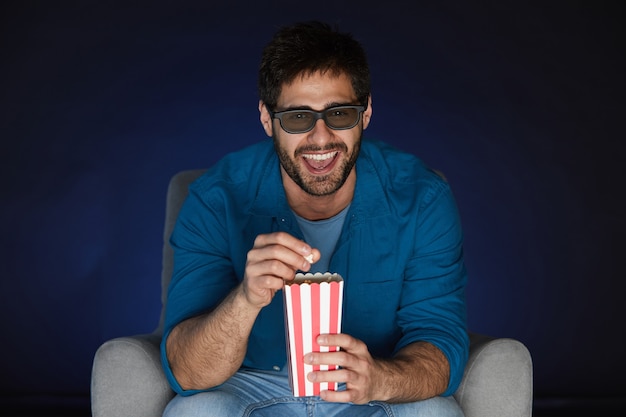 Retrato de hombre barbudo con gafas 3D comiendo palomitas de maíz mientras ve una película en casa en la oscuridad