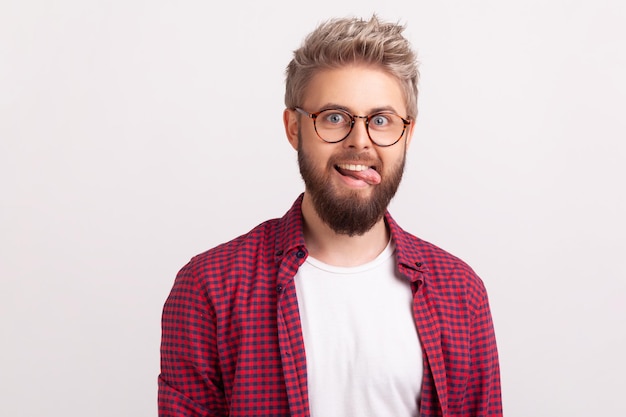 Retrato de hombre barbudo guapo loco divertido en anteojos y camisa a cuadros jugando y mostrando lengua, optimismo. Disparo de estudio interior aislado sobre fondo gris