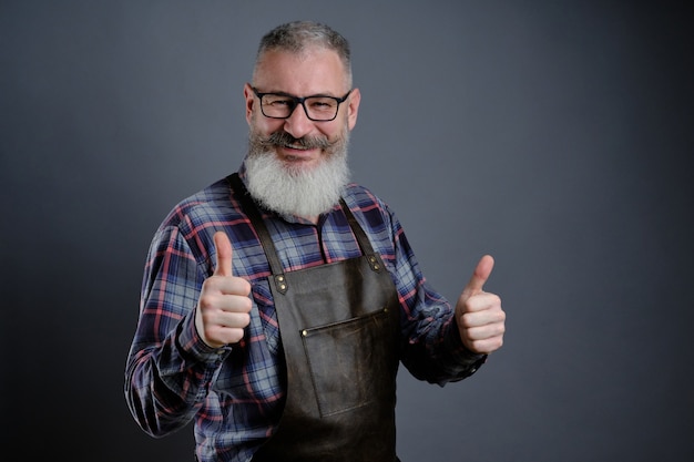 Retrato de hombre barbudo maduro guapo vestido con delantal de cuero gesticulando Thumbs up sobre pared gris trabajador caucásico con barba sonriendo