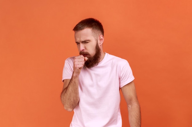 Retrato de un hombre barbudo malsano que tose y se resfría con una temperatura alta que tiene un síntoma de influenza usando una camiseta rosa Foto de estudio interior aislada en un fondo naranja