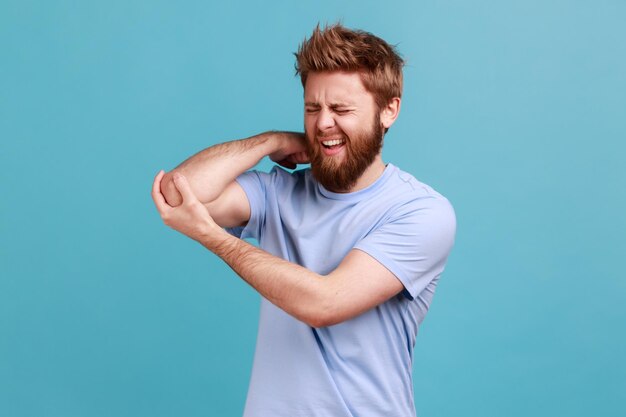 Retrato de hombre barbudo masajeando codo dolorido, frunciendo el ceño por dolor agudo, problema de tensión y tensión, nervios atrapados, artritis reumatoide. Disparo de estudio interior aislado sobre fondo azul.