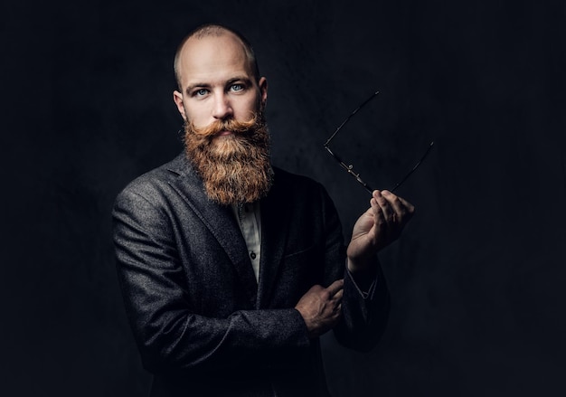 Foto retrato de hombre barbudo pelirrojo con anteojos vestido con un elegante traje de lana sobre fondo gris.