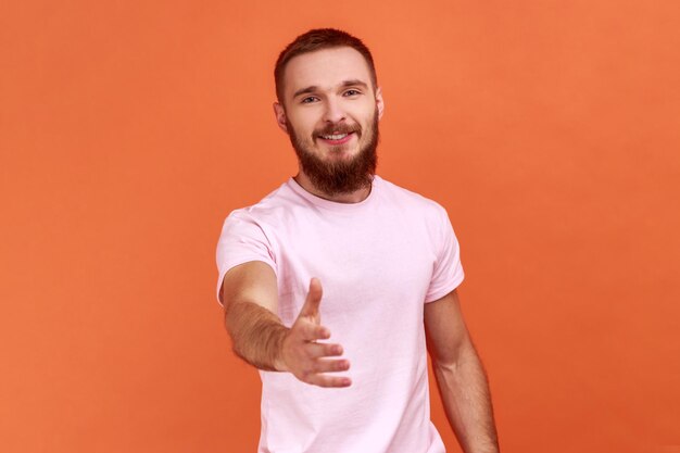 Retrato de un hombre barbudo de pie con la mano extendida, ofreciendo un apretón de manos a su pareja, saludando en una entrevista de trabajo, usando una camiseta rosa. Disparo de estudio interior aislado sobre fondo naranja.