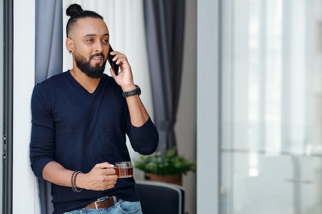 Retrato de hombre barbudo positivo bebiendo café por la mañana y hablando por teléfono con un colega o empleado