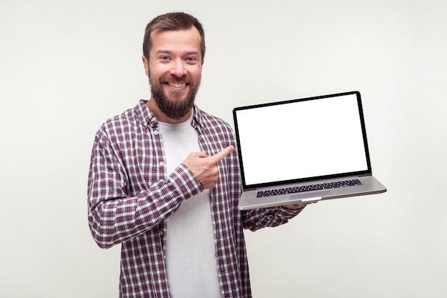 Retrato de un hombre barbudo positivo con una camisa informal a cuadros sosteniendo un portátil y señalando una pantalla vacía sonriendo satisfecho con una aplicación de ordenador en un estudio interior aislado en un lugar de fondo blanco para el anuncio