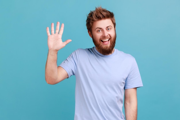 Retrato de un hombre barbudo positivo que te saluda levantando la mano y saludando, saludando, contento de verte, mirando a la cámara con una sonrisa dentuda. Disparo de estudio interior aislado sobre fondo azul.