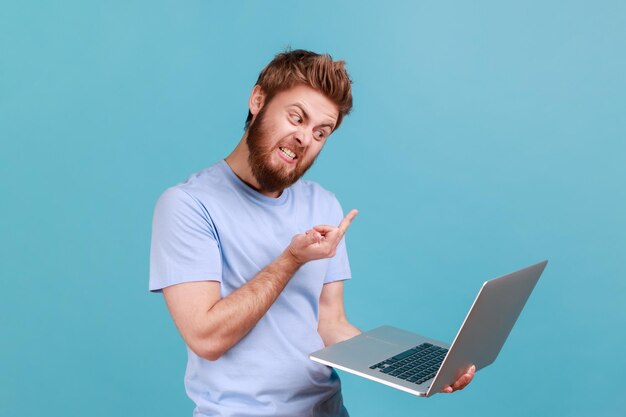 Retrato de un hombre barbudo que muestra el dedo medio a la pantalla del portátil comunicándose agresivamente en una videollamada expresando odio al trabajo o al compañero de trabajo Toma de estudio interior aislada en fondo azul