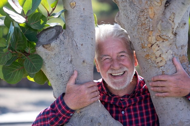 Retrato de un hombre barbudo senior disfrutando al aire libre en un parque público de pie entre dos troncos de árboles. Ancianos alegres con cabello blanco y camisa a cuadros