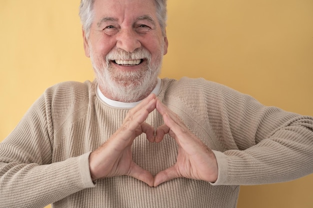 Retrato de hombre barbudo senior feliz haciendo forma de corazón con sus manos sobre fondo amarillo