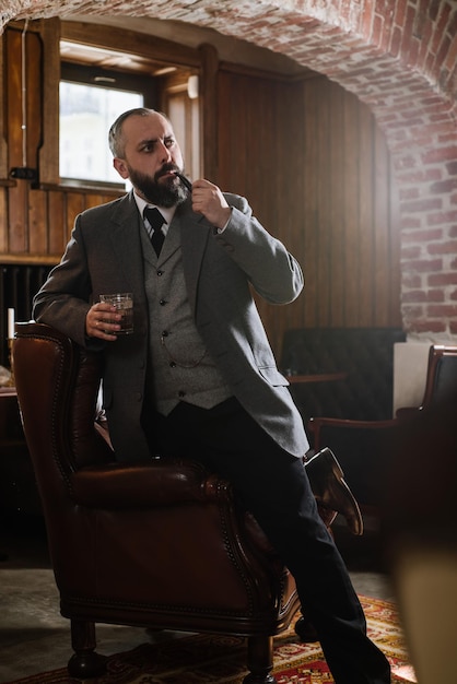 Retrato de un hombre barbudo serio con pipa sosteniendo un vaso de whisky usando traje y sentado en un gran sillón