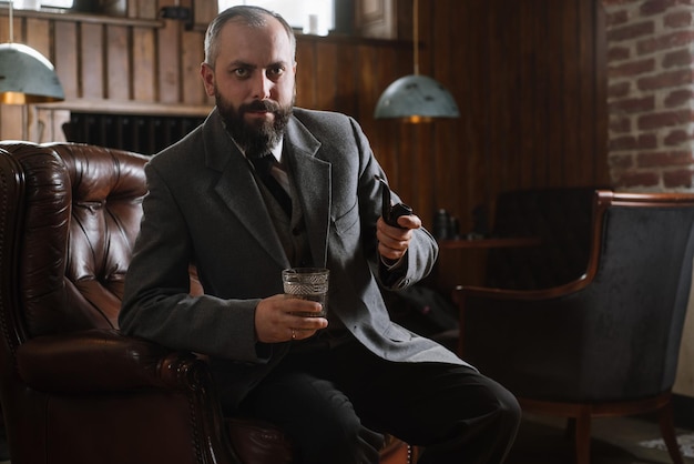 Retrato de un hombre barbudo serio con pipa sosteniendo un vaso de whisky usando traje y sentado en un gran sillón