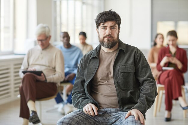 Foto retrato de hombre barbudo serio vestido con ropa casual y mirando a la cámara mientras posa en la oficina con un grupo diverso de personas en el fondo, espacio de copia