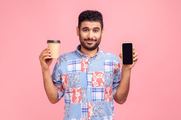 Retrato de un hombre barbudo sonriente feliz con una camisa azul de estilo casual sosteniendo una taza de café desechable y mostrando el teléfono con pantalla en blanco Foto de estudio interior aislada en fondo rosa