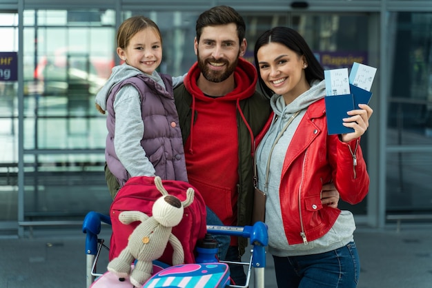 Retrato del hombre barbudo sosteniendo a su feliz hija mientras su esposa estaba cerca y mostraba boletos a la cámara. Avión de espera familiar en el concepto del aeropuerto