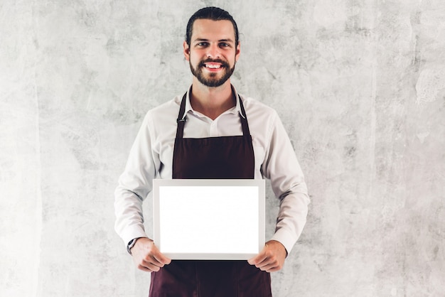 Retrato de hombre barista guapo barista hombre propietario de una pequeña empresa sonriendo y sosteniendo tablero vacío marco de madera con maqueta blanca en blanco en un café