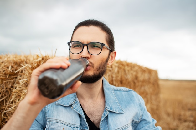 Retrato de hombre bebiendo agua de botella termo de acero cerca de un pajar.