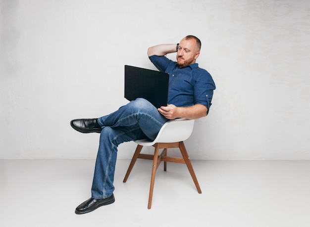 Retrato de un hombre brutal sentado en una silla contra una pared blanca con una computadora portátil y un hombre de negocios de notepada