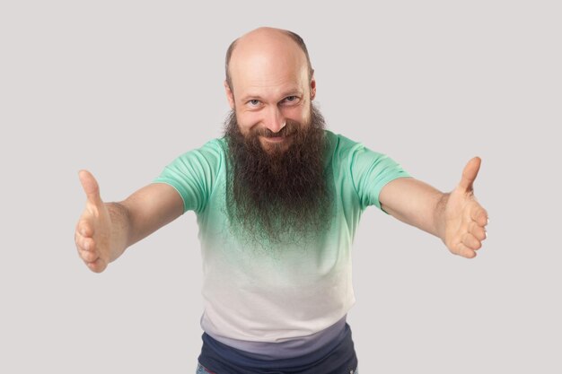 Retrato de un hombre calvo feliz de mediana edad con barba larga en una camiseta verde claro de pie con los brazos levantados, mirando con una sonrisa y quiere abrazar. tiro de estudio interior, aislado sobre fondo gris.