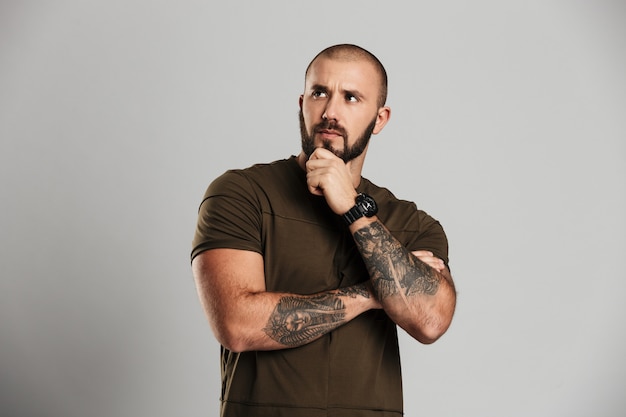 Retrato de hombre calvo varonil con tatuaje en sus brazos mirando a un lado y sosteniendo su barbilla, aislado sobre la pared gris