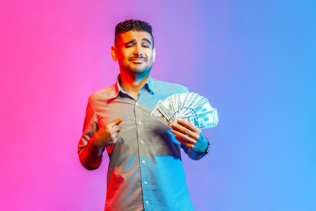 Retrato de un hombre con camisa apuntándose con el dedo a sí mismo sosteniendo un abanico de billetes de cien dólares orgulloso de su gran salario obteniendo ganancias Foto de estudio interior aislada en un colorido fondo de luz de neón