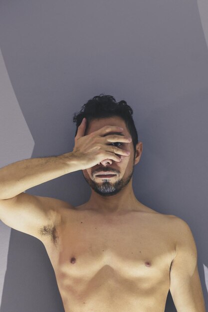 Foto retrato de un hombre sin camisa cubriendo la cara mientras está de pie contra la pared