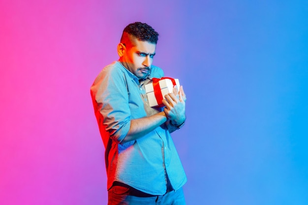Retrato de un hombre con camisa sosteniendo una caja de regalo en las manos, siendo codicioso por compartir regalos, mirando a la cámara con el ceño fruncido. Disparo de estudio interior aislado sobre fondo de luz de neón colorido.