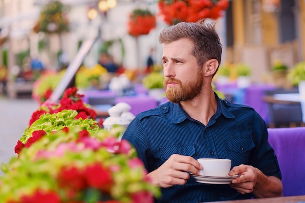 Retrato de un hombre casual pelirrojo barbudo bebe café en un café en una calle.