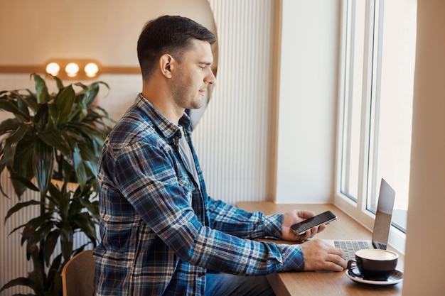 Retrato de un hombre casual sentado en una mesa cerca de la ventana, trabajando en línea con una laptop y usando el móvil