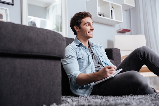 Retrato de un hombre casual sonriente tomando notas en el cuaderno mientras está sentado en la alfombra en casa