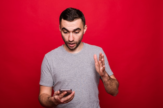 Retrato de un hombre casual sorprendido mirando teléfono móvil aislado sobre fondo rojo.