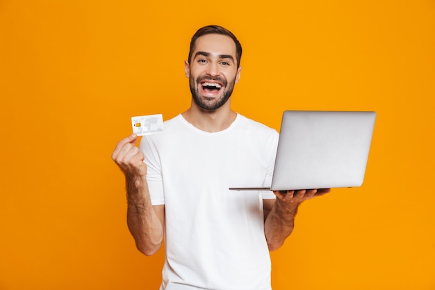 Retrato de hombre caucásico de 30 años en camiseta blanca con portátil plateado y tarjeta de crédito, aislado