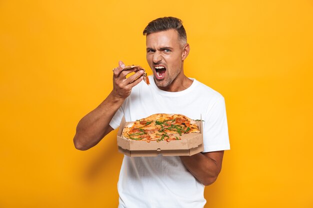 Retrato de hombre caucásico de 30 años en camiseta blanca sosteniendo y comiendo pizza mientras está de pie aislado en amarillo