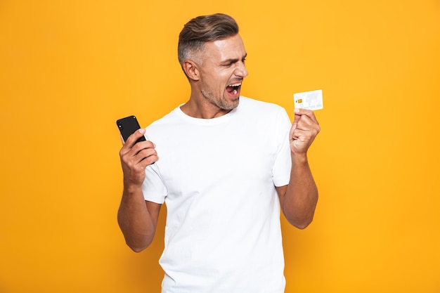 Retrato de hombre caucásico de 30 años en camiseta blanca con teléfono móvil y tarjeta de crédito aislado en amarillo