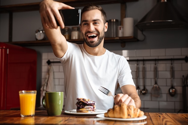 Foto retrato de hombre caucásico de 30 años tomando fotos selfie en teléfono móvil mientras desayuna en la elegante cocina de casa