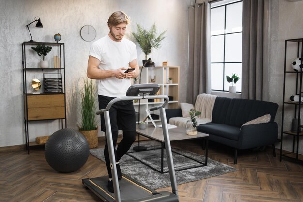 Retrato de un hombre caucásico activo en ropa deportiva usando un teléfono inteligente corriendo usando una cinta de correr