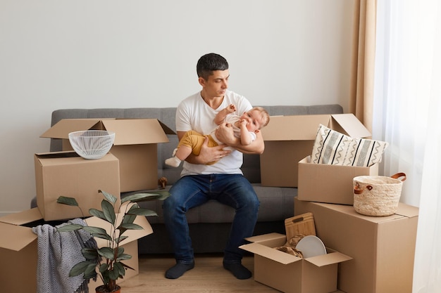 Retrato de un hombre caucásico con camiseta blanca y jeans sentado en un sofá con una hija pequeña rodeada de cajas de cartón, calmando a un bebé llorando, mudándose a un nuevo hogar con la familia.