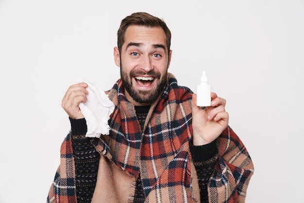 Retrato de un hombre caucásico enfermo envuelto en una manta estornudando y sosteniendo gotas nasales debido a la gripe aislado sobre la pared blanca