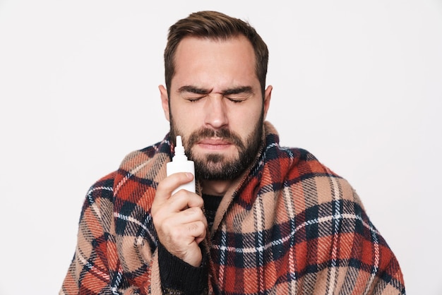 Retrato de un hombre caucásico enfermo envuelto en una manta con gotas para la nariz debido a la gripe aislado sobre la pared blanca