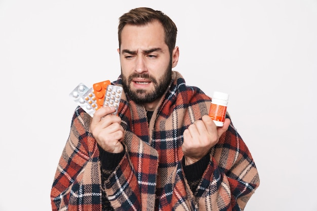 Retrato de hombre caucásico enfermo envuelto en una manta sosteniendo un montón de tabletas debido a la gripe aislado en blanco