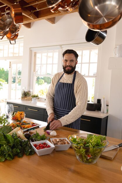 Retrato de un hombre caucásico feliz con barba cortando verduras con delantal en la cocina. Concepto de vida doméstica, tiempo libre, salud y felicidad.