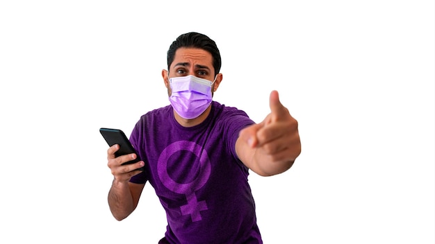 Retrato de un hombre caucásico feminista que usa una camisa morada con un símbolo de género femenino mientras señala con la mano la cámara y usa un teléfono inteligente con máscara médica durante el brote del virus de la corona.