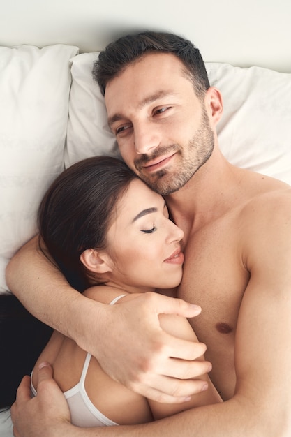Foto retrato de un hombre caucásico joven guapo de barba oscura complacido abrazando a su esposa dormida en la cama