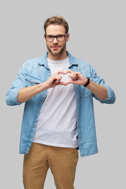 Retrato de hombre caucásico joven guapo haciendo signo de corazón con las manos sobre gris