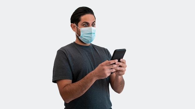 Retrato de hombre caucásico levantando las cejas debido a la sorpresa con el teléfono inteligente y la máscara médica durante el brote del virus corona. Persona de reacción sorprendida con dispositivo de teléfono sobre fondo blanco.