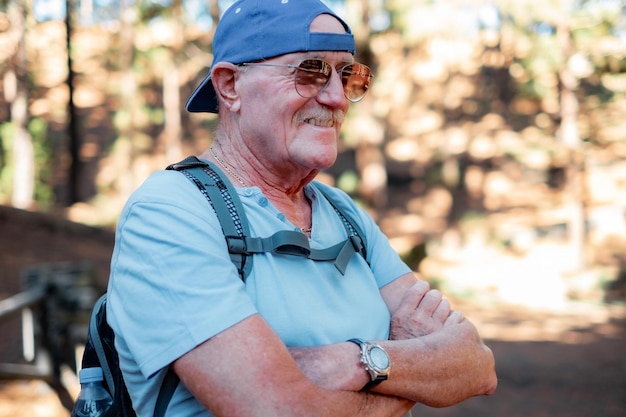 Retrato de un hombre caucásico mayor feliz con los brazos cruzados de pie en el bosque listo para una excursión a la montaña Hombre jubilado con sombrero, gafas de sol y mochila