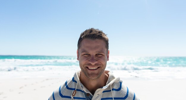 Retrato de un hombre caucásico de pie en la playa con el cielo azul y el mar en el fondo, sonriendo a la cámara