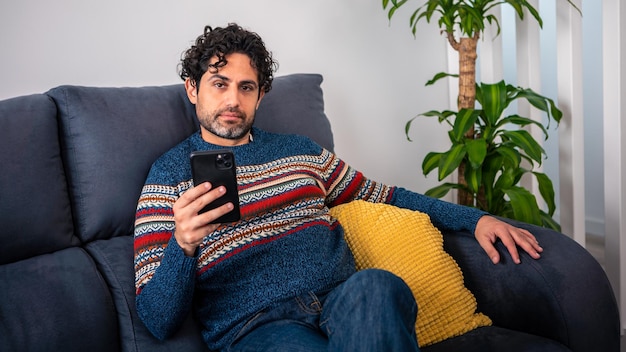 Retrato de hombre caucásico con smartphone sentado en el sofá en el interior de la hermosa casa. El hombre caucásico comprueba el dispositivo del teléfono en el sofá de la casa.